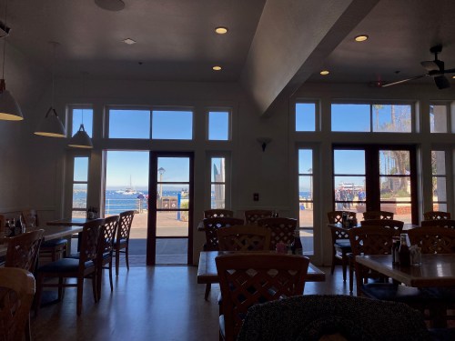 Interior of a restaurant with wooden tables and chairs, featuring large windows overlooking a waterfront view.