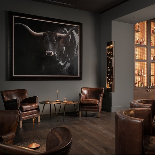 A stylish lounge with leather chairs, a bull painting, and a well-stocked bar against a dark wall.