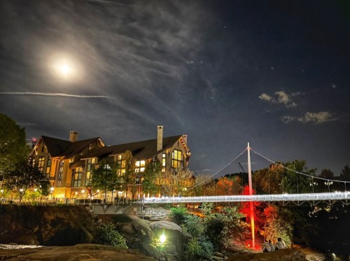 A moonlit scene featuring a large lodge beside a river, connected by a bridge, with trees and clouds in the background.