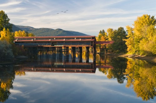 A serene river scene with a wooden structure reflecting in the water, surrounded by autumn trees and a clear sky.