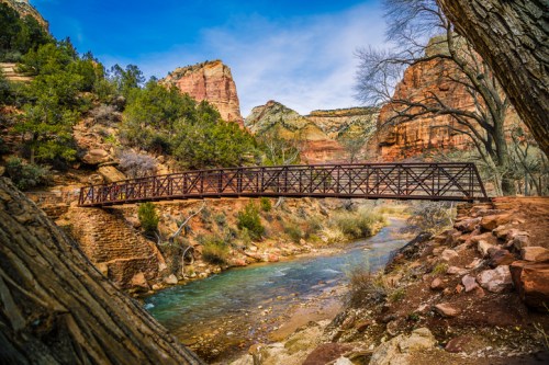 A metal bridge spans a river in a scenic canyon, surrounded by lush greenery and towering red rock formations.