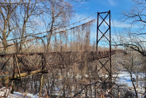 An old, rusted suspension bridge stretches over a snowy landscape, surrounded by bare trees and a clear blue sky.