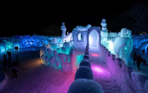 A colorful ice castle illuminated at night, with glowing blue and purple lights and visitors exploring the icy structures.