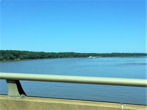 A calm river scene with green trees lining the shore and a barge in the distance under a clear blue sky.