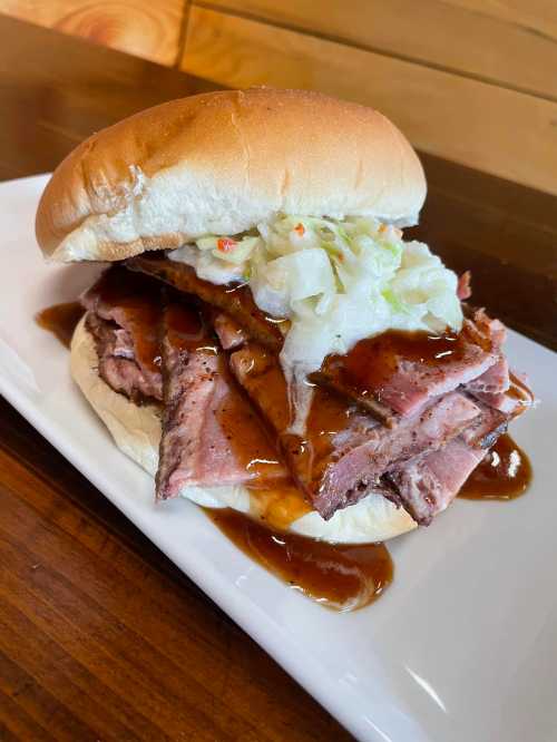 A hearty sandwich with layers of beef, topped with coleslaw and drizzled with barbecue sauce, served on a bun.