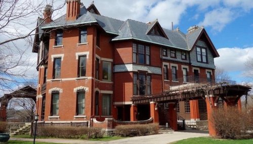 A large, historic red brick mansion with a steep roof and multiple windows, surrounded by greenery and blue skies.
