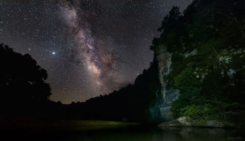 A starry night sky with the Milky Way over a dark, wooded landscape and a calm river reflecting the stars.
