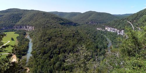 A panoramic view of lush green hills and a winding river surrounded by dense forests under a clear blue sky.