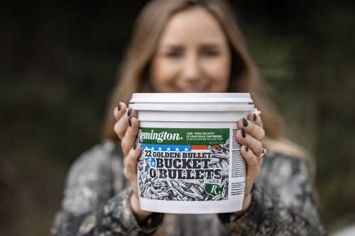 A person holds a bucket of Remington .22 Golden Bullet ammunition, smiling in a natural outdoor setting.