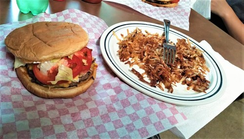 A hamburger with lettuce and tomato next to a plate of crispy hash browns on a checkered tablecloth.