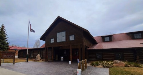 A log cabin-style building with a red roof, flagpole, and landscaped entrance on a cloudy day.