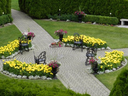 A circular garden with yellow flowers, black butterfly-shaped chairs, and neatly arranged greenery along a stone path.