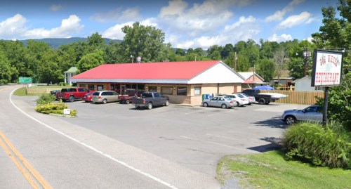 A roadside restaurant with a red roof, surrounded by trees and parked cars, under a blue sky with clouds.