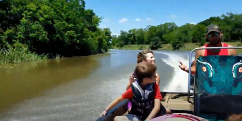 A man and two children enjoy a boat ride on a river, surrounded by lush greenery and a clear blue sky.