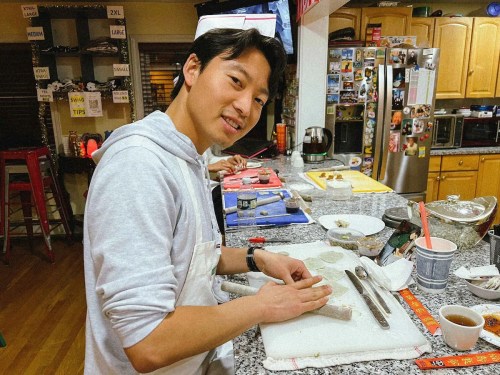 A person in a kitchen prepares food, smiling while working on a cutting board with various kitchen tools around.