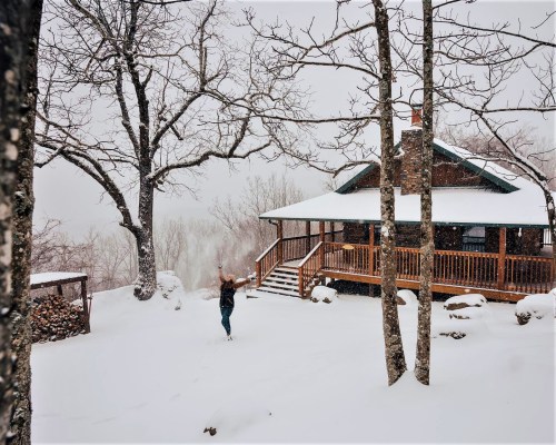 A person joyfully plays in the snow outside a cozy cabin surrounded by trees on a snowy day.