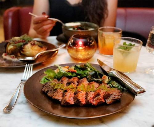A plate of sliced steak with greens, surrounded by drinks and a candlelit table setting.