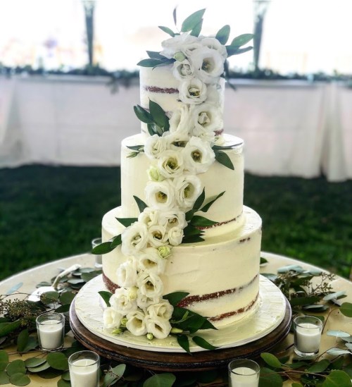 A three-tiered wedding cake adorned with white flowers and greenery, set on a rustic wooden stand.
