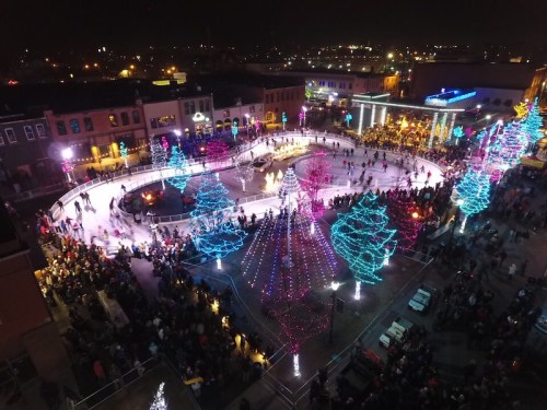 Aerial view of a festive ice skating rink surrounded by colorful lights and a large crowd enjoying the winter scene.