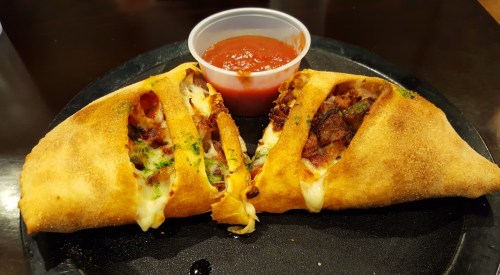 A golden-brown calzone filled with meat and vegetables, served with a side of marinara sauce.