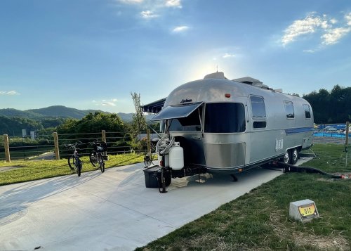 A silver Airstream trailer parked on a concrete pad, with bicycles nearby and mountains in the background under a blue sky.