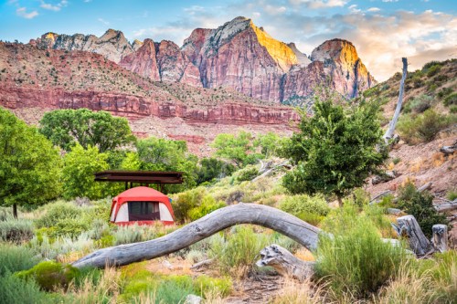 A red tent set in a lush valley with towering mountains and colorful rock formations in the background.