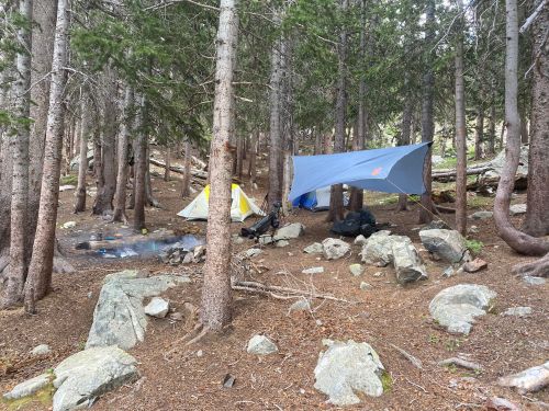 Two tents and a tarp set up in a forest clearing, surrounded by trees and rocks. A campfire area is visible.