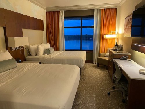 A hotel room with two beds, a desk, and a window showing a view of water at dusk. Warm lighting and modern decor.