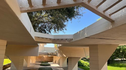 A modern architectural structure with open spaces, featuring angular lines and greenery in the background.