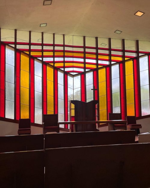 Interior of a church featuring vibrant stained glass windows in red and yellow, with a wooden pulpit and seating.