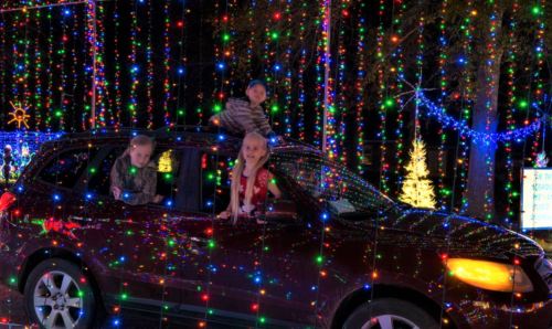 Three children peek out of a car decorated with colorful holiday lights, surrounded by a festive light display.