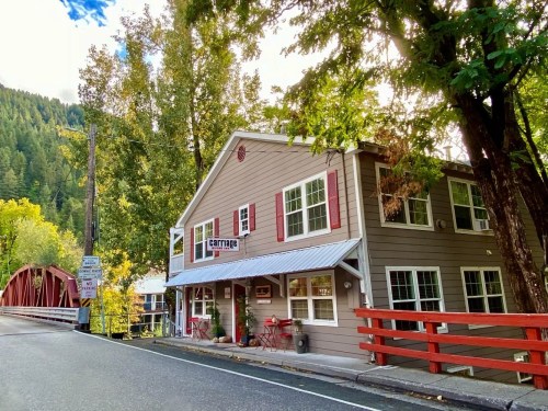 A charming two-story building with red shutters, located near a tree-lined road and a red bridge in a scenic area.