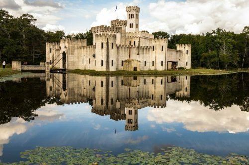 A castle with towers reflected in a calm lake, surrounded by trees and a cloudy sky.
