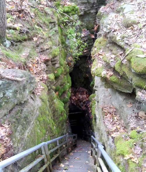 A narrow, moss-covered stone pathway leads into a dark, wooded cave entrance surrounded by greenery and fallen leaves.