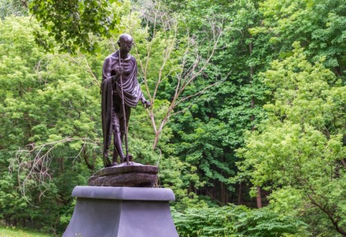 A bronze statue of Gandhi stands on a pedestal, surrounded by lush green trees in a serene outdoor setting.