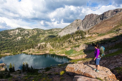 A hiker in a purple shirt stands on a rocky ledge overlooking a serene mountain lake surrounded by lush greenery.