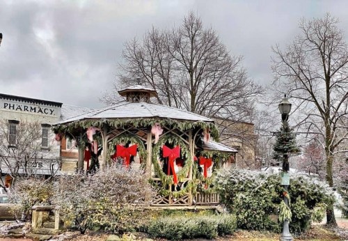 A decorated gazebo in a snowy park, adorned with red bows and greenery, surrounded by bare trees and buildings.