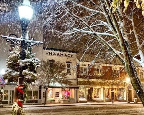 A snowy street scene at night featuring a pharmacy and charming shops, illuminated by a streetlamp.
