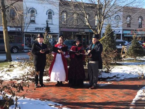 Four people in historical costumes sing in a snowy town square, with shops and trees in the background.