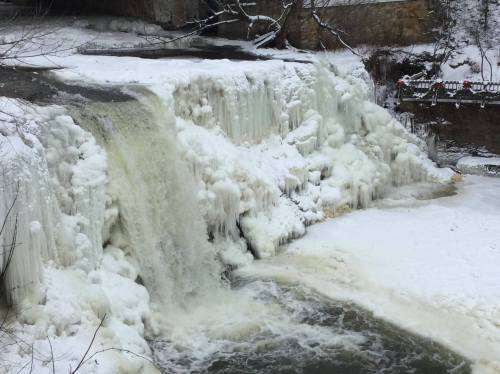 A frozen waterfall cascading over icy cliffs, surrounded by snow-covered trees and a partially frozen river below.
