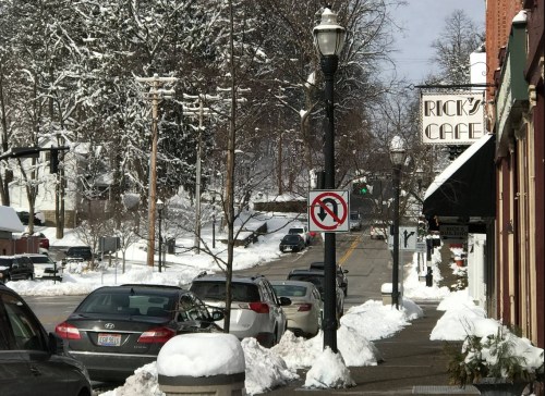 A snowy street scene featuring parked cars, a café sign, and a no parking sign amidst snow-covered trees.