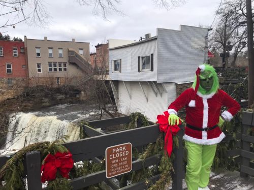 A person dressed as the Grinch stands by a closed park sign near a waterfall, decorated with holiday garlands.