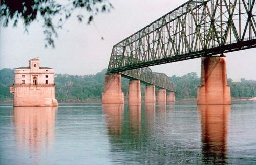 A historic bridge spans a river, with a small building on the shore reflecting in the calm water below.