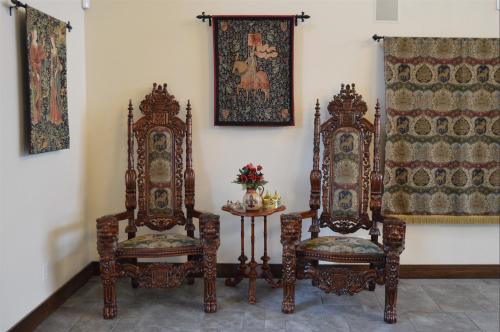 Two ornate wooden chairs with decorative cushions, flanked by wall hangings and a small table with a floral arrangement.