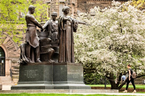 A bronze statue featuring three figures, surrounded by blooming trees and a grassy area, with a historic building in the background.