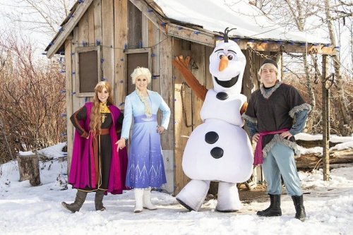 Four characters from a winter scene stand outside a wooden cabin, surrounded by snow: two princesses, a snowman, and a man.