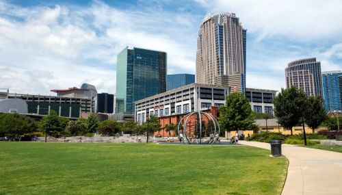 A city park with green grass and trees, featuring modern skyscrapers and a sculpture in the background under a blue sky.
