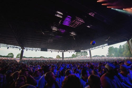 A large crowd at an outdoor concert venue, with a beach ball flying above the audience under colorful stage lights.