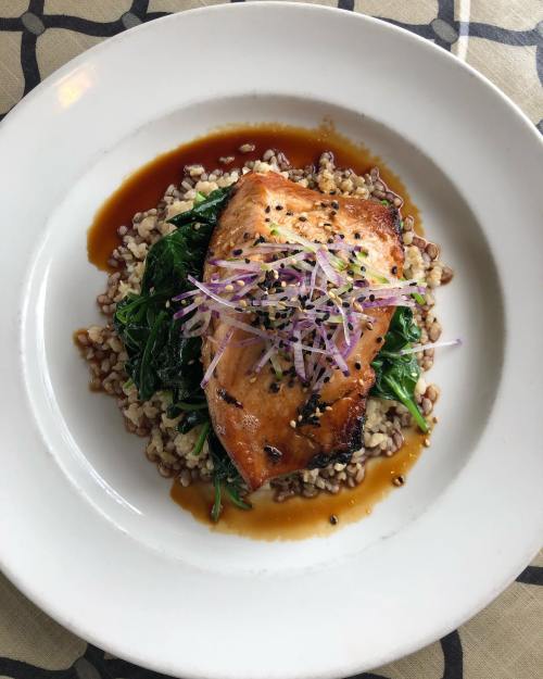 A plate of grilled salmon on a bed of grains and sautéed greens, garnished with sprouts and a drizzle of sauce.