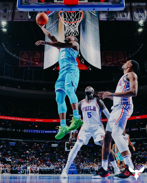 A player in a teal uniform leaps to dunk a basketball while two defenders watch in a packed arena.